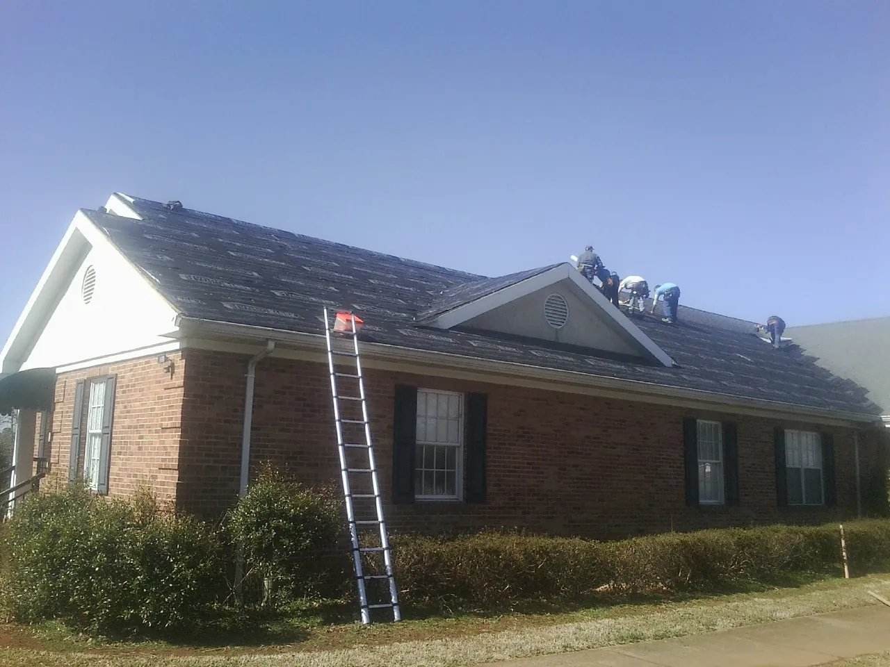 Crew tearing shingles off a brick ranch roof with a ladder set — Emergency Roof Repair work by our crew in Amlin