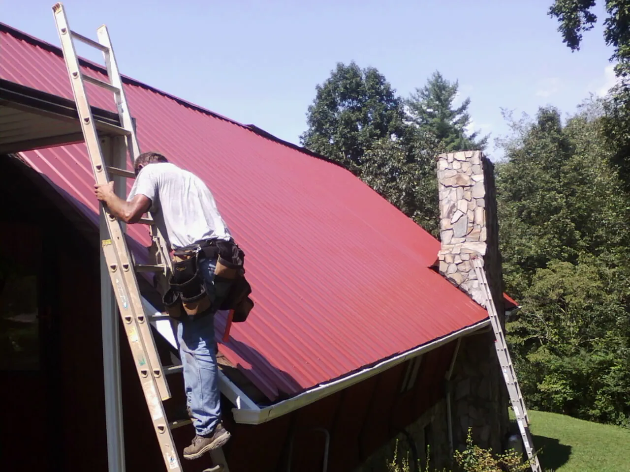 Roofer on a ladder beside a red metal roof and stone chimney — Soffit & Fascia Repair by Roofing Brothers in Amlin